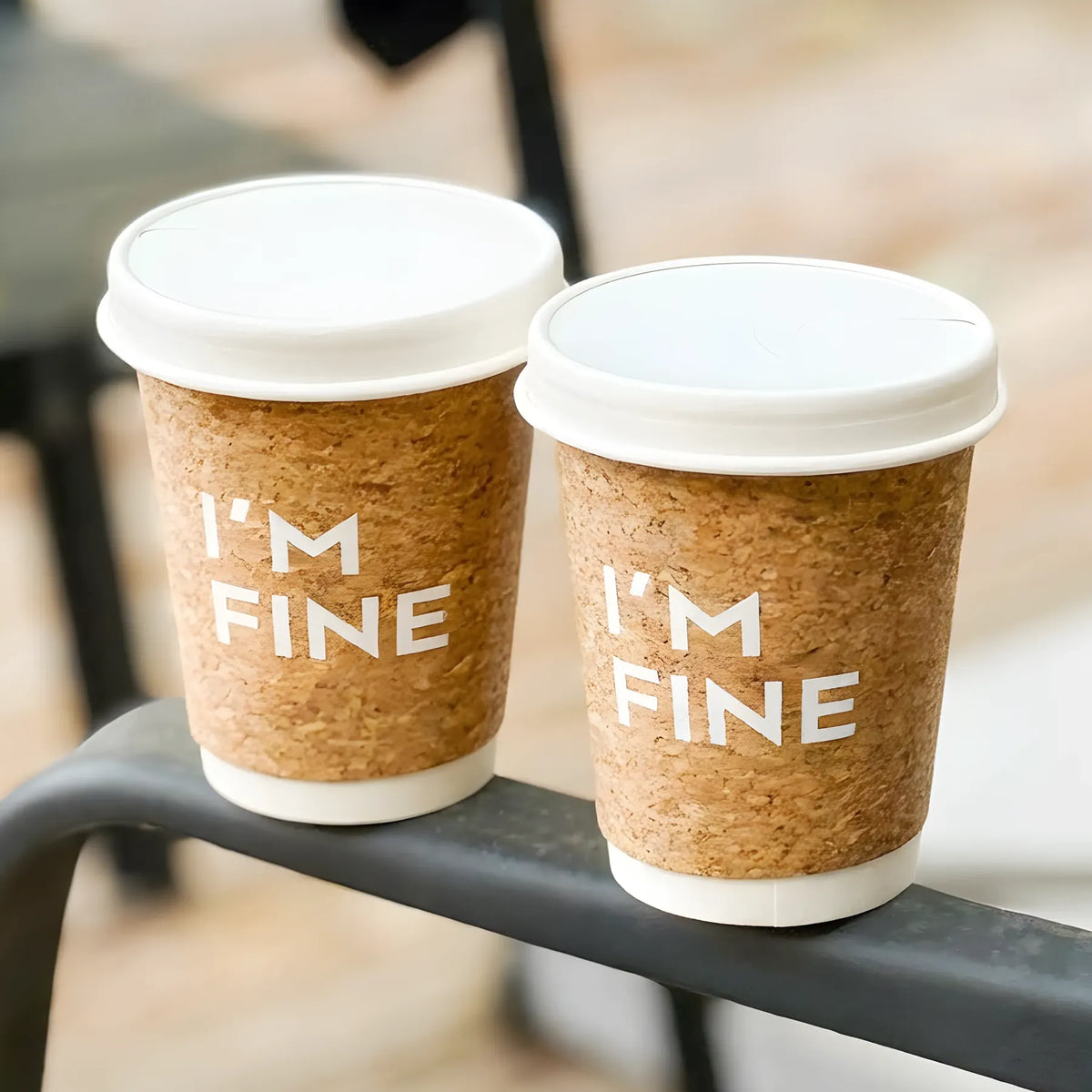 two coffee cups with a cork finish and text that says, "I'm fine" sit on a railing.
